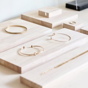 Puede incluir: Un conjunto de expositores de madera con pulseras y collares de oro. Los expositores son de madera clara y están dispuestos en forma escalonada.
