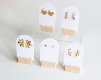 May include: Six pairs of earrings displayed on white card stands with wooden bases. The earrings are gold and silver with various designs, including a flower, a crescent moon, and a fan.