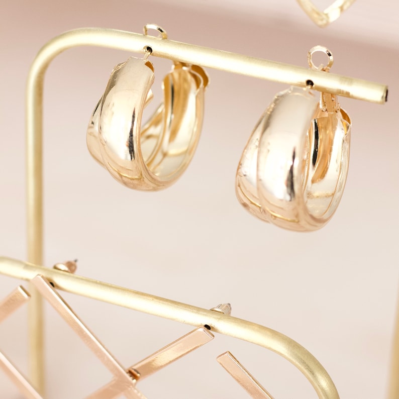 Op de afbeelding: Twee gouden creolen met een dik, afgerond ontwerp. De oorbellen worden gepresenteerd op een gouden metalen standaard.