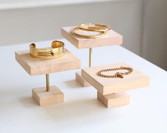 May include: Three wooden display stands with gold jewellery on them. The stands are square and have a small post in the centre. The jewellery includes a bracelet, a pair of hoop earrings, and a chain bracelet.