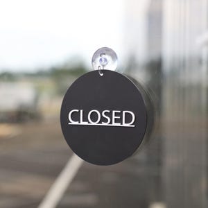 Op de afbeelding: Een zwarte, ronde bord met het woord "CLOSED" in witte letters. Het bord is met een transparante zuignap aan een glazen oppervlak bevestigd. De wazige achtergrond suggereert een buitenomgeving.