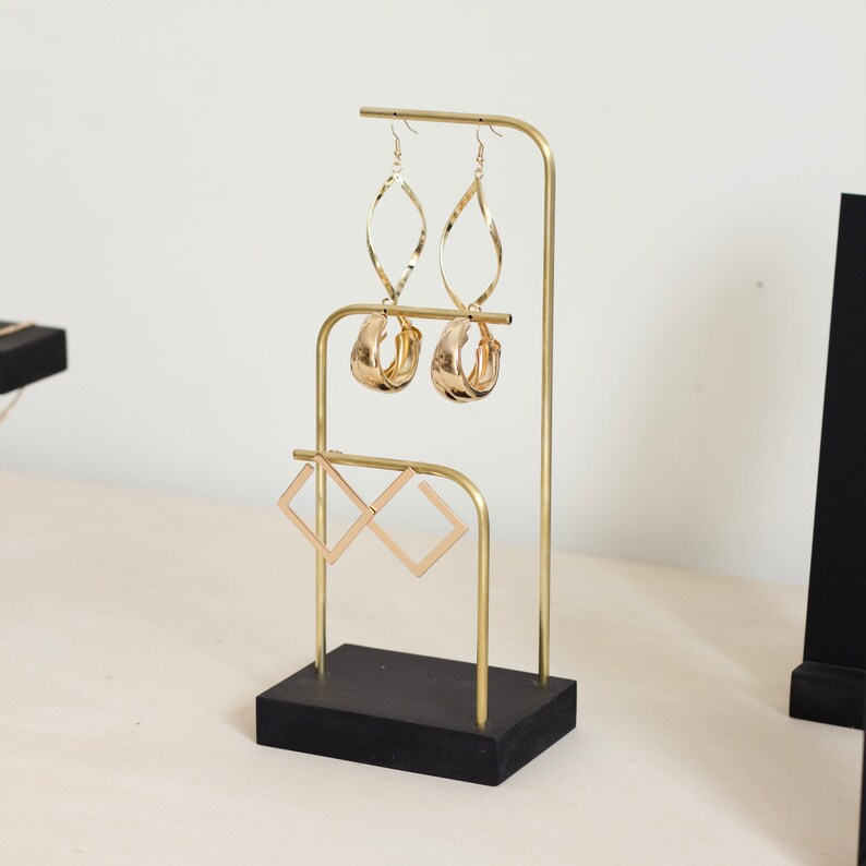 Op de afbeelding: Gouden metalen oorbellenstandaard met drie niveaus. De standaard heeft een zwarte houten basis en beschikt over een verscheidenheid aan gouden oorbellen.
