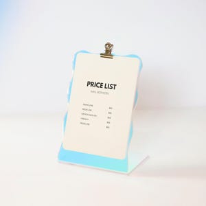May include: A white price list sign with a blue iridescent frame and a silver clip. The sign lists prices for manicure, pedicure, design add-on, and french pedicure.