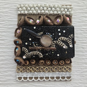 May include: A mixed media art piece featuring a black rectangular box with a starry night design, adorned with acorn caps, decorative trim, and iridescent gems. The artwork incorporates a feather, a circular element, and zebra-like patterns.