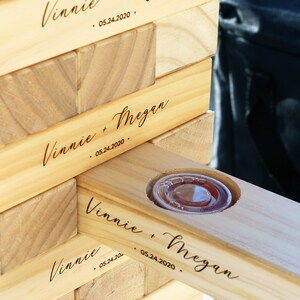 Puede incluir: Un juego de Jenga de madera con los nombres "Vinnie + Megan" y la fecha "05.24.2020" grabados en los bloques. Un bloque tiene un recorte circular que contiene un recipiente de pl&aacute;stico transparente.