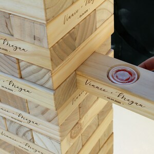 Puede incluir: Un juego de apilar bloques de madera con los nombres "Vinnie & Megan" y la fecha "05.24.2020" grabados en los bloques. Un bloque tiene un peque&ntilde;o recipiente de pl&aacute;stico transparente con l&iacute;quido rojo. El juego est&aacute; parcialmente montado.