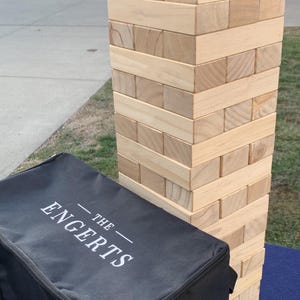 May include: A large wooden block stacking game, with a black carrying case. The game is made of natural wood blocks, stacked to form a tower. The black case has the text "THE ENGERTS" printed on it.