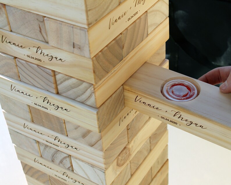 Puede incluir: Un juego de apilar bloques de madera con los nombres "Vinnie & Megan" y la fecha "05.24.2020" grabados en los bloques. Un bloque tiene un peque&ntilde;o recipiente de pl&aacute;stico transparente con l&iacute;quido rojo. El juego est&aacute; parcialmente montado.