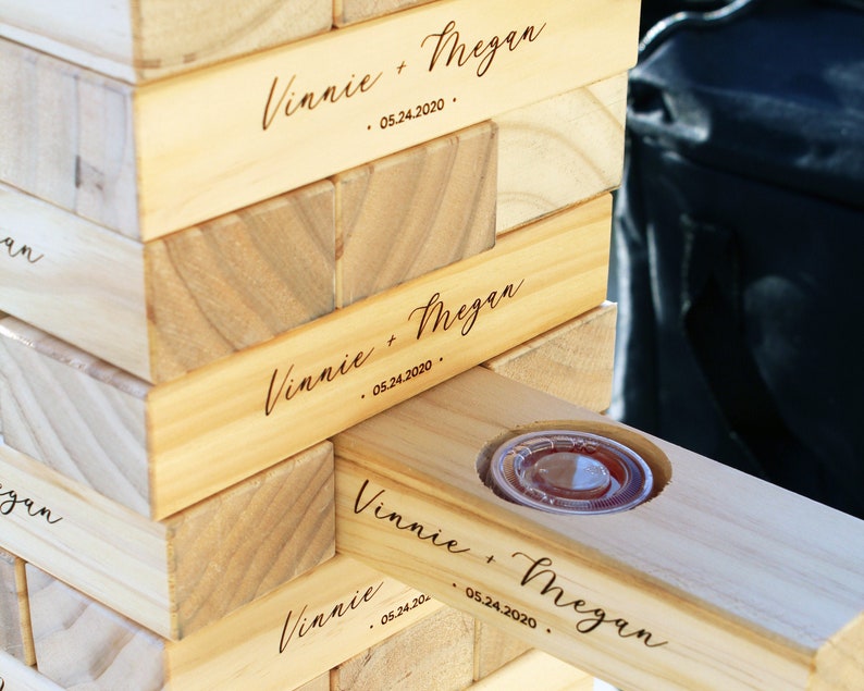 Puede incluir: Un juego de Jenga de madera con los nombres "Vinnie + Megan" y la fecha "05.24.2020" grabados en los bloques. Un bloque tiene un recorte circular que contiene un recipiente de pl&aacute;stico transparente.