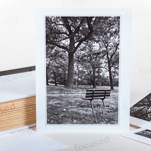 May include: A black and white photograph of a wooden bench in a park setting. The bench is surrounded by trees and grass. The photo is framed in a white mat.