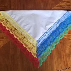 Puede incluir: Un pañuelo triangular blanco con ribete de encaje colorido. El ribete de encaje es rojo, amarillo, azul y verde. El pañuelo está hecho de una tela blanca suave y está colocado sobre una superficie de madera.