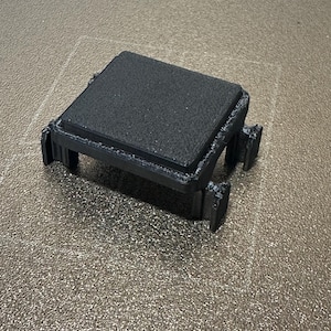 May include: A black, 3D-printed square object with a textured surface and four supporting legs. The top surface appears to be a platform, and the object is sitting on a textured, gray surface.