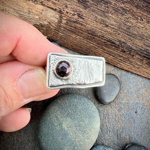 May include: A silver ring with a rectangular face and a textured surface. A small, round, dark red gemstone is set in the center of the ring. The ring is displayed against a wooden background with several gray stones.