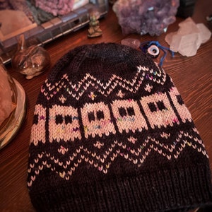 Puede incluir: Un gorro de punto marrón oscuro con un patrón geométrico blanco y rosa. El patrón presenta una serie de cuadrados pequeños con un contorno blanco y un interior rosa.