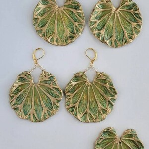 Pu&ograve; includere: Un set di quattro orecchini a forma di foglia verde e oro. Gli orecchini sono in metallo e hanno una superficie testurizzata. Gli orecchini sono appesi a catene d'argento.