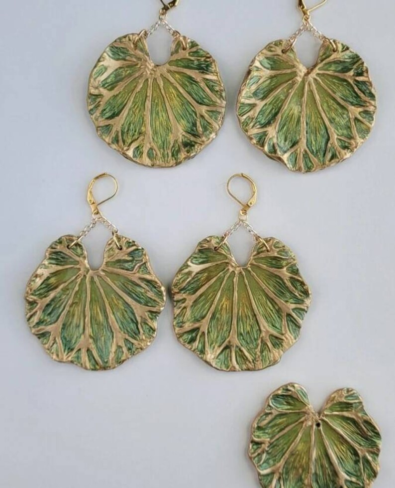 Pu&ograve; includere: Un set di quattro orecchini a forma di foglia verde e oro. Gli orecchini sono in metallo e hanno una superficie testurizzata. Gli orecchini sono appesi a catene d'argento.