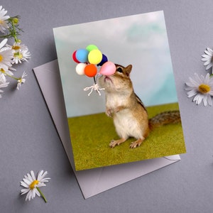 May include: A cute chipmunk holding a bunch of colorful balloons. The chipmunk is standing on a green grassy surface with a blue sky in the background.
