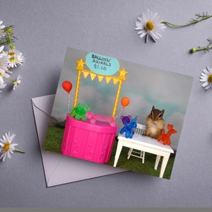 May include: A chipmunk sits at a miniature balloon animal stand with a sign that reads "Balloon Animals $1.00". The stand is pink with a yellow awning and has a variety of colorful balloon animals on display.