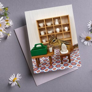 May include: A greeting card featuring a cartoon illustration of a chipmunk sitting at a miniature table with a cup of coffee and a toolbox. The chipmunk is wearing a white shirt and a blue tie. The background is a tiled floor with a miniature shelf filled with white ceramic items.