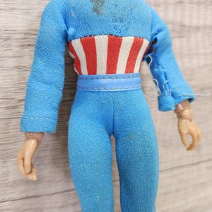 May include: A blue and red action figure with a blue belt and a red, white, and blue striped shirt. The figure has a torn sleeve and is missing a hand.