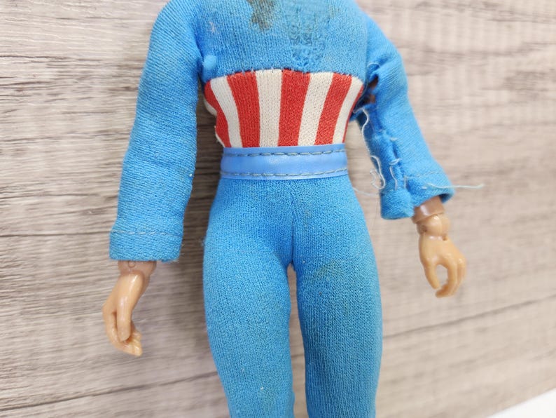 May include: A blue and red action figure with a blue belt and a red, white, and blue striped shirt. The figure has a torn sleeve and is missing a hand.