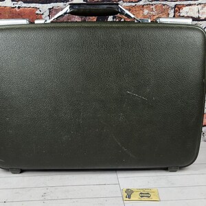 May include: A rectangular olive green hard-shell suitcase with a textured exterior finish. The suitcase features a black handle with metallic silver accents at the top and rounded corners, presenting a structured design for storage and transport.
