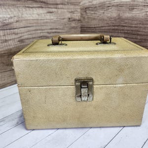 May include: A vintage beige cardboard box with a brown plastic handle and a metal latch. The box has a textured surface and is in good condition.