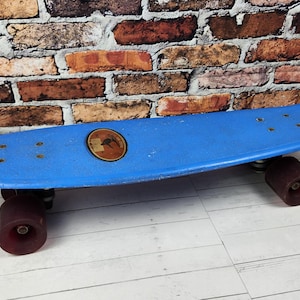 May include: A vintage blue skateboard with dark red wheels. The skateboard has a round emblem in the center and is set against a brick wall and a wooden floor.