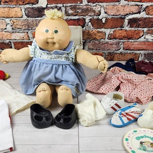 May include: A vintage Cabbage Patch Kids doll with various outfits and accessories. The doll is wearing a blue dress and black shoes. Other items include shorts, shirts, shoes, a comb, and a plate.