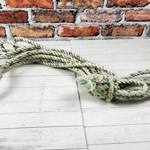 May include: A coiled rope with a white and green pattern. The rope is lying on a white wooden surface, with a brick wall in the background. The rope is knotted at one end.