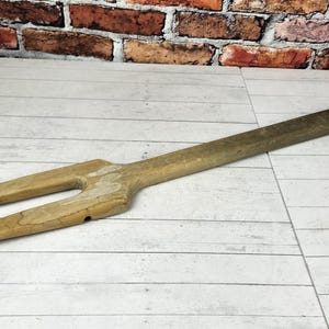 May include: A vintage wooden fork-like tool with a long handle and two prongs. The tool is made of light-colored wood and shows signs of wear. The background features a brick wall and a white wooden surface.