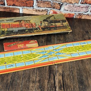 May include: Vintage toy train set with a colorful track, a box labeled "Automatic Loader," and a red box with the number "297." The track is orange, blue, and yellow, with a detailed design. The box features a train illustration.
