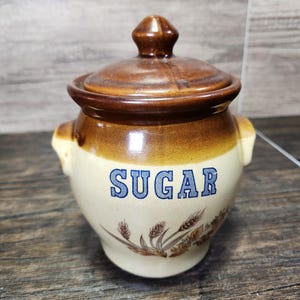 May include: A brown and cream ceramic sugar jar with a lid. The jar has a blue and white floral design and the word "SUGAR" written in blue letters.