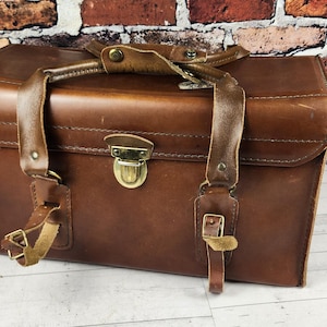 May include: A vintage brown leather camera bag with a top handle and shoulder straps. The bag has a rectangular shape with a metal clasp closure and brass hardware. The bag is sitting on a white wooden surface.