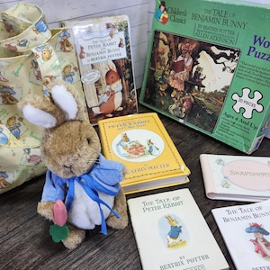 May include: A collection of Peter Rabbit themed items including a tote bag, a plush toy, a book, and a puzzle. The tote bag is yellow with a white pattern of Peter Rabbit characters. The plush toy is a brown rabbit with blue clothing. The book is yellow with a picture of Peter Rabbit on the cover. The puzzle is a wooden puzzle with a picture of Peter Rabbit on the box. The text on the puzzle box reads "The Tale of Benjamin Bunny by Beatrix Potter".