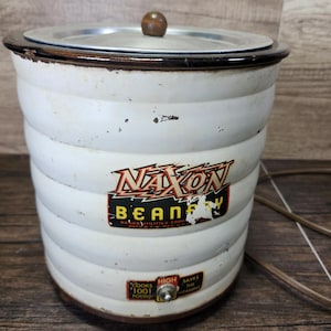 May include: White vintage Naxon Beanery electric slow cooker with a brown lid and a cord. The cooker has a white and red logo that says "Naxon Beanery".
