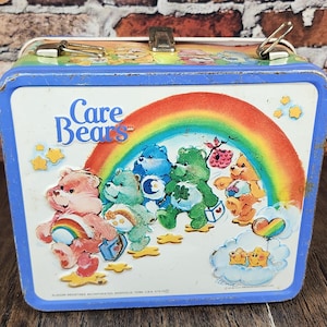 May include: Vintage Care Bears lunchbox with a blue frame and handle. The front features a rainbow and several Care Bears characters. The lunchbox is made of metal and has a clasp closure. The Care Bears logo is visible.