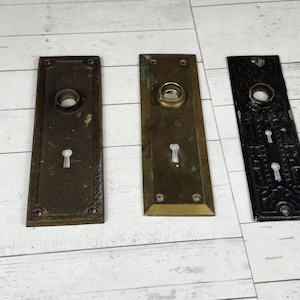 May include: Four antique door hardware pieces. Includes a white, ornate door plate, and three rectangular plates in bronze, brass, and black. Each plate has a keyhole and a circular opening for the doorknob.