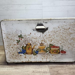 May include: Vintage white metal bread box with a rustic design featuring a teapot, oil lamp, and fruit. The box has a handle on the top and is slightly rusted.
