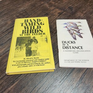 May include: Two vintage books on a wooden surface. One, titled "HAND-TAMING WILD BIRDS AT THE FEEDER," is yellow with black text and a photo. The other, "DUCKS AT A DISTANCE," is white with a duck illustration and text.
