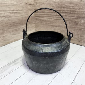 May include: A dark gray cast iron cauldron with a curved handle. The cauldron has a wide, open mouth and a rounded body. The handle is made of a dark metal and is attached to the sides of the cauldron. The cauldron is sitting on a white wooden surface.