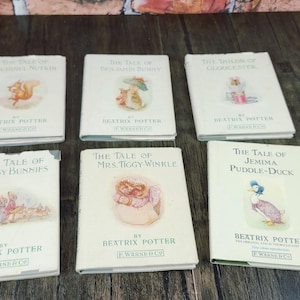 May include: Six vintage Beatrix Potter children's books are arranged on a dark wooden surface. The book titles include "The Tale of Squirrel Nutkin," "The Tale of Jemima Puddle-Duck," and "The Tale of Mrs. Tiggy-Winkle."