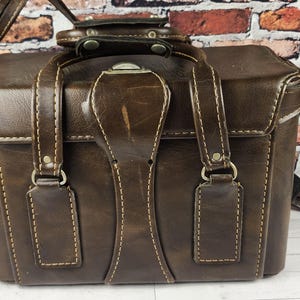 May include: A vintage brown leather camera bag with a shoulder strap and top handle. The bag features a rectangular shape with stitched detailing and metal hardware. The leather has a worn appearance, suggesting age and use.