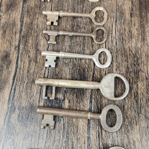 May include: A collection of seven antique keys of varying sizes and designs, arranged vertically on a wooden surface. The keys are made of metal, with visible wear and tear, suggesting age and use. The background is a dark wood grain.