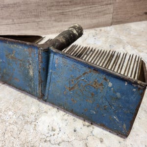 May include: A blue metal rake with a wooden handle. The rake has a scoop on one end and a row of metal teeth on the other.