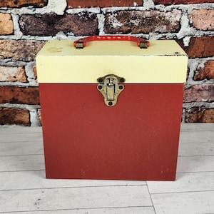 May include: A vintage red and cream-colored storage box with a red handle and a metal clasp. The box is rectangular and sits on a light wood surface in front of a brick wall.