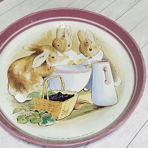 May include: Round metal tray with a pink rim and a painted illustration of three bunnies gathered around a bowl. A basket of berries and a pitcher are also depicted. The tray has a vintage, storybook aesthetic.