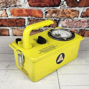 May include: A yellow vintage radiation survey meter with a handle, dial, and a circular logo. The device has a rectangular shape and is made of metal. The text on the device reads "MODEL RM-18-2A THE VICTOREEN INSTRUMENT CO. CLEVELAND, OHIO".