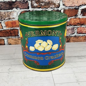 May include: A cylindrical green tin with a matching lid. The tin features the word "VERMONT" in yellow, a blue banner with crackers, and the words "Cinnamon Crackers". The tin is on a white surface.