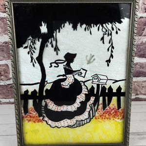May include: Framed silhouette art featuring a woman in a long dress, holding a book, under a tree. The black silhouette is set against a white background with a yellow and orange foreground, framed in a silver-colored metal.